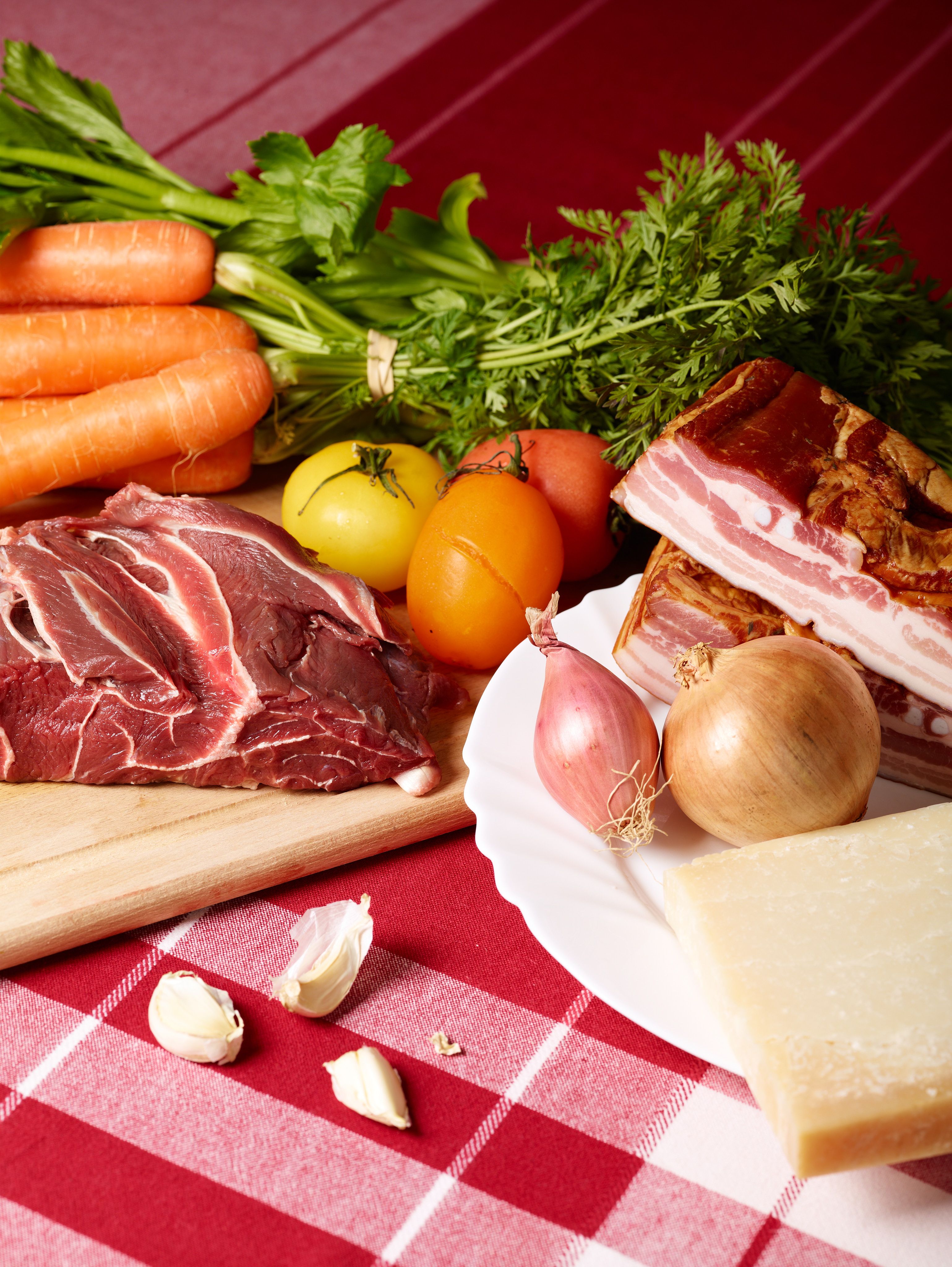 Still life of beef, tomatoes, carrots, garlic, onions, ham and cheese on a red tablecloth